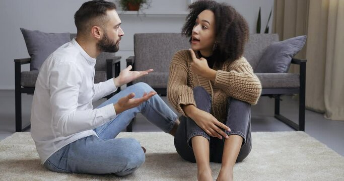 Multiethnic young couple sitting on floor near couch sofa curly woman wife and caucasian husband man quarreling shouting outraged together sort out relationship, separation divorce problem concept