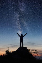 Fototapeta na wymiar Vertical shot of space traveler standing on rocky hill with night beautiful sky and Milky way on background. Silhouette of cosmonaut in space suit looking at stars and spreading arms out to sides.