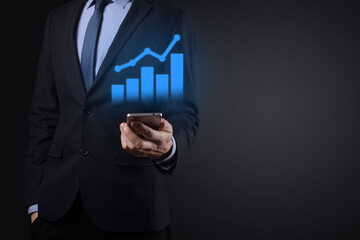 Businessman holding tablet and showing a growing virtual hologram of statistics, graph and chart with arrow up on dark background. Stock market. Business growth, planing and strategy concept.