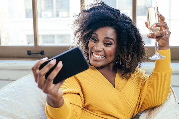 Portrait of a smiling  woman celebrating with Champagne glass on a phone video call at home on couch