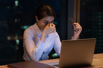 Tired Indian female employee sit in office late hours take off glasses overwhelmed with computer...