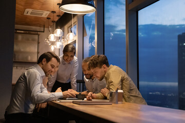 Smiling diverse businesspeople gather in office late hours watch video on smartphone together. Happy multiracial colleagues coworkers work relax after day using cellphone at workplace at night.