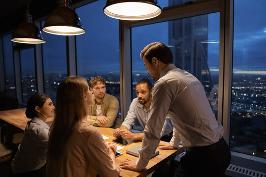 Multiracial businesspeople gather at team meeting in office at night, meet deadline. Diverse multiethnic colleagues work late hours, discuss company business project together. Teamwork concept.