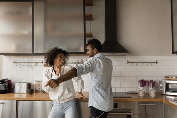 Excited young African American couple have fun enjoy early morning in modern renovated kitchen. Happy millennial biracial man and woman dance celebrate moving relocation to own home. Realty concept.