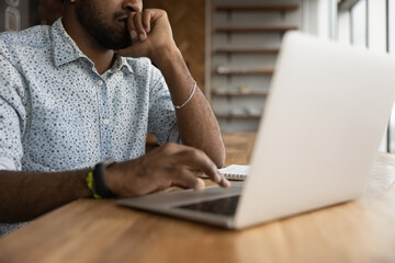 Crop close up of pensive African American man sit at desk in home office look at laptop screen working online. Biracial male busy using modern computer gadget, study distant or take online course.