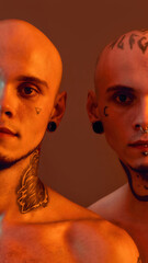 Cropped portrait of young half naked twin brothers with tattoos and piercings looking at camera, posing together isolated over orange background