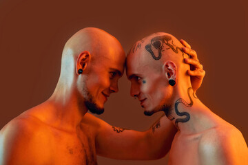 Portrait of young twin brothers with tattoos and piercings standing head to head, smiling at each other isolated over orange background