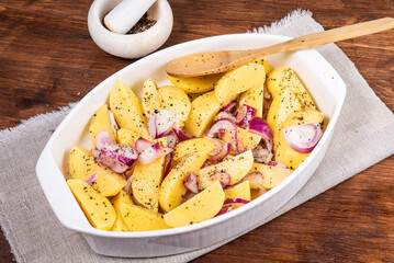 Sliced raw potatoes with onions with black pepper and sausages in a baking dish on a wooden table, top view