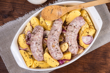 Sliced raw potatoes with onions with black pepper and sausages in a baking dish on a wooden table, top view
