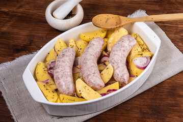 Sliced raw potatoes with onions with black pepper and sausages in a baking dish on a wooden table, top view