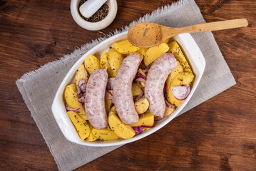 Sliced raw potatoes with onions with black pepper and sausages in a baking dish on a wooden table, top view