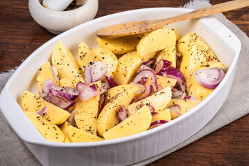 Sliced raw potatoes with onions with black pepper and sausages in a baking dish on a wooden table, top view, close-up