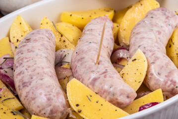 Sliced raw potatoes with onions with black pepper and sausages in a baking dish on a wooden table, top view, close-up