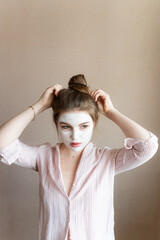 Fototapeta premium a girl in a white clay mask stands by the wall straightens her hair in a bun