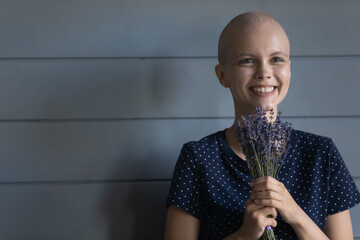 Head shot close up smiling hairless woman cancer patient holding flowers, standing on grey wooden...