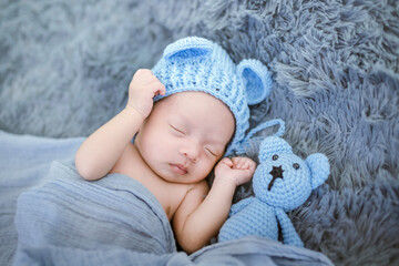 Portrait of twenty seven day old newborn asian baby boy in knitted blue hat with teddy bear sleeping on blue fur bed in the studio