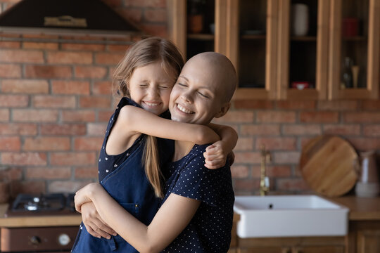 Close up happy sick hairless mother cancer patient and little daughter cuddling, standing in modern kitchen at home, loving smiling adorable child girl and mum hugging, enjoying tender moment