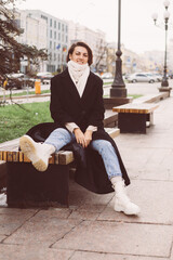 Outdoor portrait of caucasian woman wearing black winter coat and scarf sitting on bench, city on background.
 