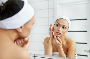 Young lady looks in bathroom mirror touching moisturized soft healthy sensitive skin . Morning...