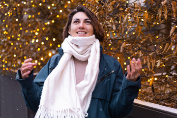 Caucasian happy smiling woman enjoying snow and winter, wearing warm scarf,  christmas lights on background, woman waiting for christmas miracle, street frame 2021    