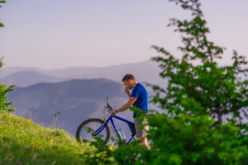 Tired cyclist is wiping his sweat off his face while pushing his bicycle uphill on a dirt road in a...