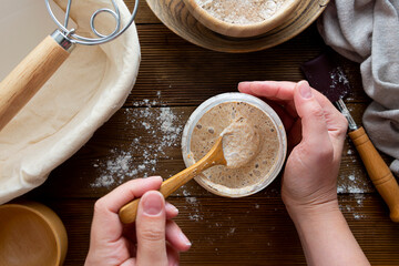 Starter sourdough. Female hands holding a jar with sourdough over wooden background.
