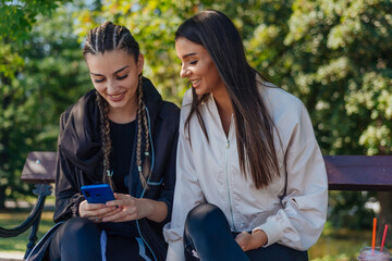 Two young girls are sitting on a bench outdoor while one of them is showing something to her friend...