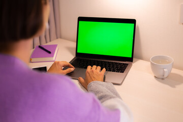 Woman at home in bedroom  watching green screen on laptop on table, notebook and pen lying near by. 