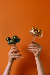 Two champagne glass with colorful Christmas bauble balls in women's hands against red wall. Creative minimalistic Christmas / New Year holidays celebration composition.