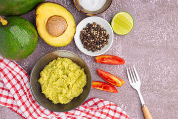 Organic avocado with seed, avocado halves, whole fruit, and fresh guacamole in a bowl with salt and pepper on a small dish on gray stone background. Top view. Concept of healthy fruit