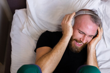 Happy caucasian man on bed wearing headphone listen enjoy his favorite music, resting alone, dancing.  