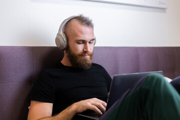 Caucasian bearded man in headphones in bedroom on bed working on laptop from home,  typing, thinking.  