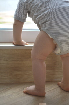 Close up of baby in grey body trying to stand with support on a wooden floor. Little feet and legs. Baby holding step. Play. Child learning to stand.