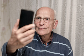 Mature hoary man holds black smartphone in hand and has facetime with friends.