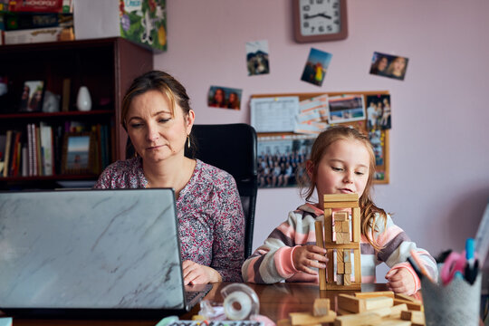 Woman mother working doing her job remotely during video chat call stream online course webinar on laptop from home while her daughter playing with bricks toy. Woman sitting at desk