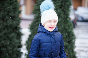 a girl in a blue jacket and a white hat is playing in a snowy yard. sincere joy of the child from playing in the fresh air. raising immunity in winter. playing snowballs in the winter forest
