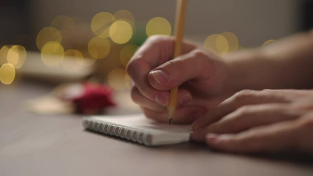 Slow motion orbit shot of man writes christmas gifts in notepad with a pencil under warm light in the evening