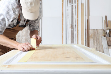 Male carpenter working the wood in carpentry workshop, putting paper masking tape on a wooden door,...