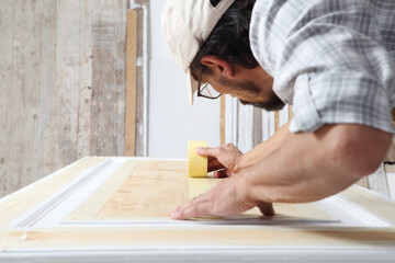 Male carpenter working the wood in carpentry workshop, putting paper masking tape on a wooden door, wearing overall and cap