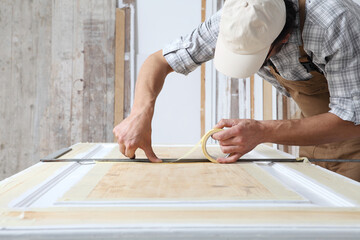 Male carpenter working the wood in carpentry workshop, putting paper masking tape on wooden door following the line of metal square ruler, wearing overall and cap
