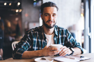 Portrait of happy male writer with textbook smiling at camera spending daytime for creating idea in cafe interior, successful hipster guy with coffee cup sitting at cafeteria table and posing