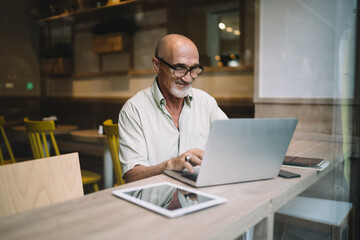 Mature Caucasian man in spectacles typing on laptop computer share content in blog browsing web pages and information, successful male freelancer doing remote job using netbook in coworking space