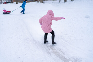 A young woman in a pink down jacket falls on a slippery ice path in a city park.