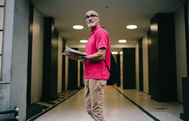 Contemplative male tourist in optical eyewear for vision protection thoughtful looking away during travel sightseeing near urban building, pensive aged man in classic glasses holding location map