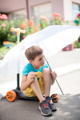Cheerful boy in a blue t-shirt sits on a skateboard under a white umbrella