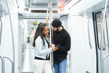 Portrait of a young interracial couple in the subway car standing, laughing at something they see on the mobile. Public transportation.