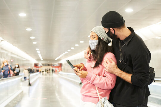 A Young Interracial Couple Of Lovers With Masks And Wool Hats In A Subway Corridor Or At The Airport Laughing And Consulting Something Funny On The Smart Phone.