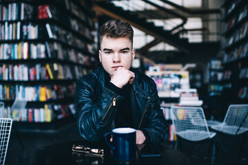 Serious man sitting at table in bookstore and touching chin