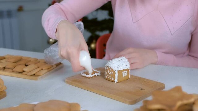 Woman is decorating gingerbread house with sugar sweet icing, hands closeup. Cooking, baking and decorating homemade gingerbread house for Christmas holidays. New Year family traditions.