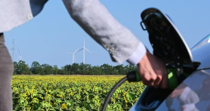A man connects an electric car to a charger and several wind generators stop in the background. Overload in the electrical network due to electric cars.